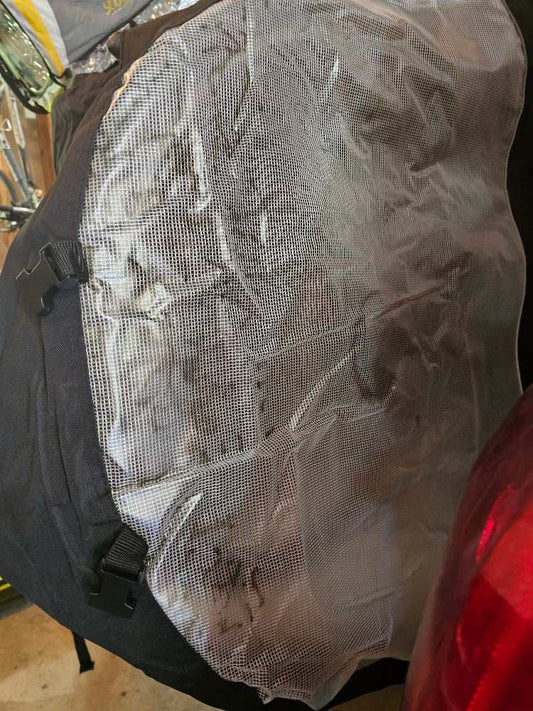 A close-up of a heavy-duty black backpack partially covered with a crumpled, see-through sheet designed for UV protection. The sheet obscures some of the contents of the backpack and is part of the Cosmetic Defect - Quad Bike Rack Cover For Transport (Fits 3-4 Bikes) Extra Large Translucent Ends by Formosa Covers. A red object, likely a car, is visible on the right side of the image.