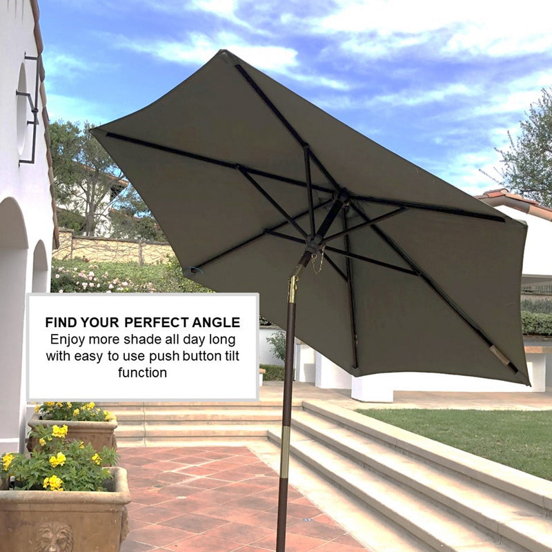 A 7ft Wooden Patio Garden Market Umbrella with Tilt Mechanism by Formosa Covers, featuring a sage green canopy and wooden crank and tilt, is angled to the right, providing shade on a sunny day with a partly cloudy sky. A sign next to the umbrella reads, "FIND YOUR PERFECT ANGLE - Enjoy more shade all day long with an easy-to-use push button tilt function.