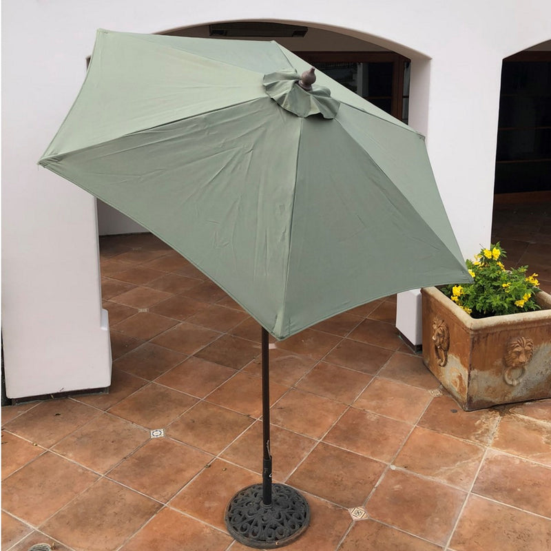 A Formosa Covers 7ft Wooden Patio Garden Market Umbrella with Tilt Mechanism in Sage Green is open and standing on a tiled floor. The umbrella features a wooden pole with a decorative base. In the background, there is a white arched wall and a flowerpot with yellow flowers. The UV-treated canopy ensures protection from the sun's rays while you enjoy your patio space.