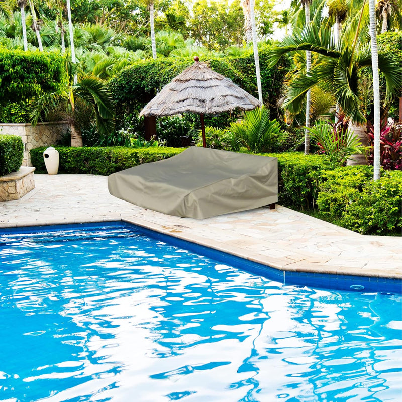 A serene backyard features a swimming pool bordered by lush greenery and stone pavers. Two lounge chairs, protected by the Patio Double Chaise Lounge Cover 84"L x 60"W x 24"H in Classic Taupe from Formosa Covers, sit under a thatched umbrella on the patio near the pool. The atmosphere is tranquil and tropical, enhanced by weatherproof patio covers.