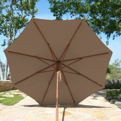 A large 9ft Market Patio Umbrella with an 8 Rib Replacement Canopy in Carmel Latte from Formosa Covers stands on a stone-paved area. Green trees surround the space, providing shade and a serene outdoor setting.