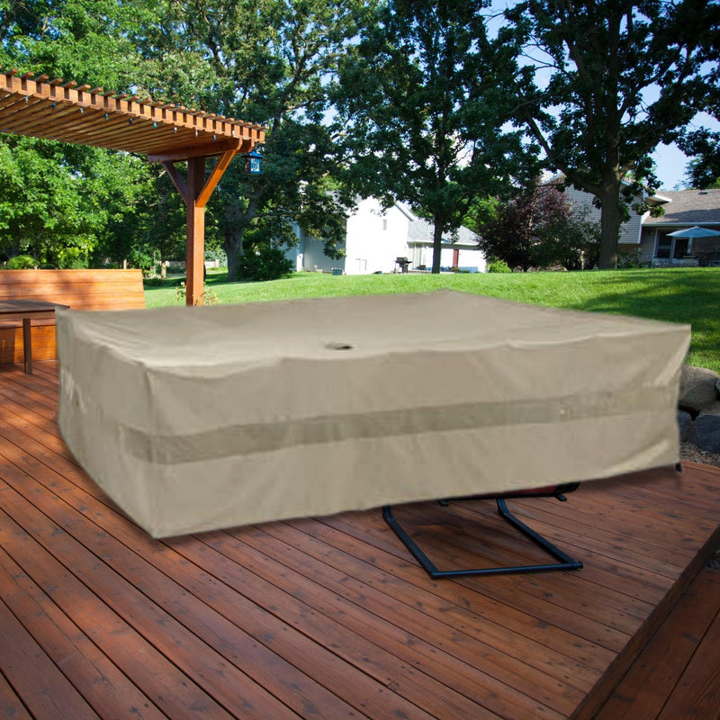 A large rectangular table on a wooden deck is covered with a Formosa Covers Patio Set Cover for Rectangular or Oval Tables, measuring 120"L x 86"W x 45"H in Classic Taupe. The deck is partially shaded by a wooden pergola, and there is lush green foliage and a white house visible in the background.