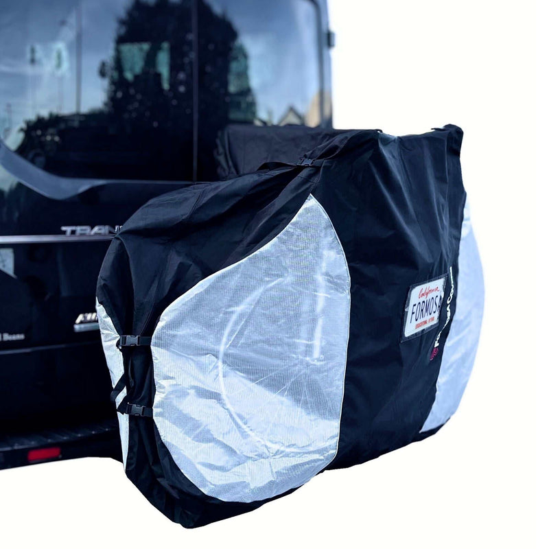 A large, black waterproof Dual Bike Rack Cover for E-Bike Transport (Fits 1-2 E-Bikes), branded "Formosa Covers," is securely attached to the rear of a vehicle using a hitch mount bike rack. The cover, featuring large translucent ends, protects the bikes from UV and water damage while allowing their outlines to remain partially visible against the backdrop of the vehicle's rear and partial reflections.