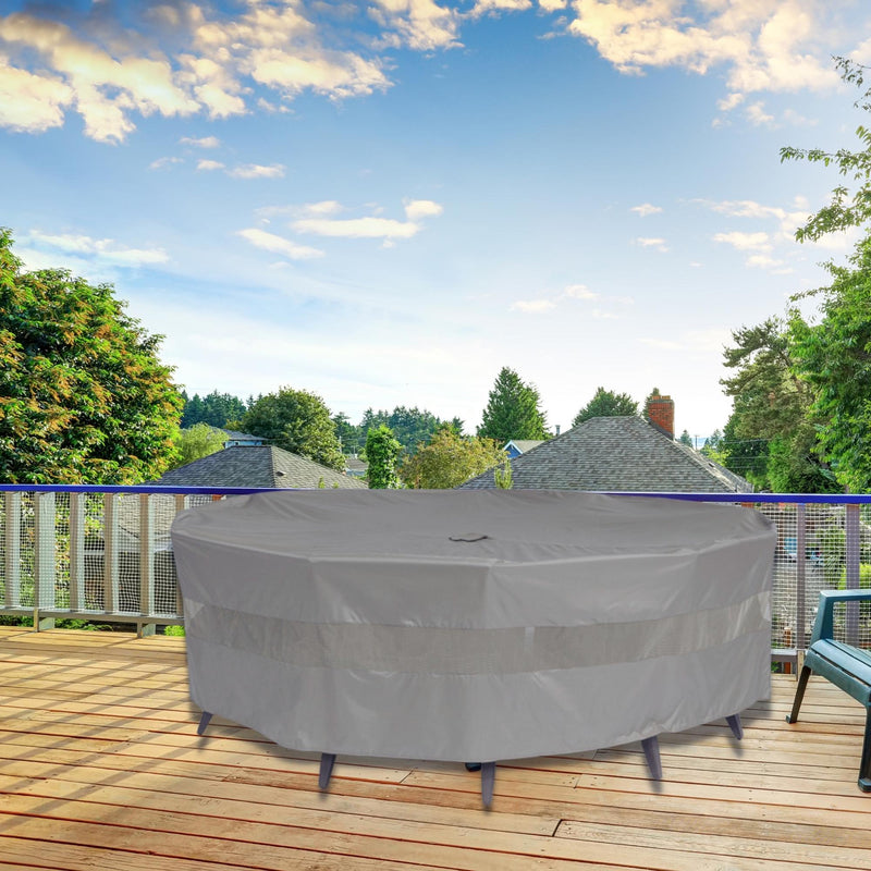 A round outdoor furniture set, protected by the Formosa Covers Patio Conversation Set Cover in Reserve Grey (104"Dia. X 31"H with Umbrella Hole), stands on a wooden deck. The deck is enclosed by a white fence, with lush green trees visible in the background under a partly cloudy sky. Two chairs are positioned on the right side.