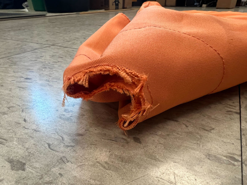 A close-up of a damaged section of the "Cut Finial Hole Open Box - 9ft Market Patio Umbrella 8 Rib Replacement Canopy Tangerine" from Formosa Covers, showing frayed edges and a hole on one end. The durable polyester fabric is laid out on a tiled floor, with boxes and other items visible in the blurred background.