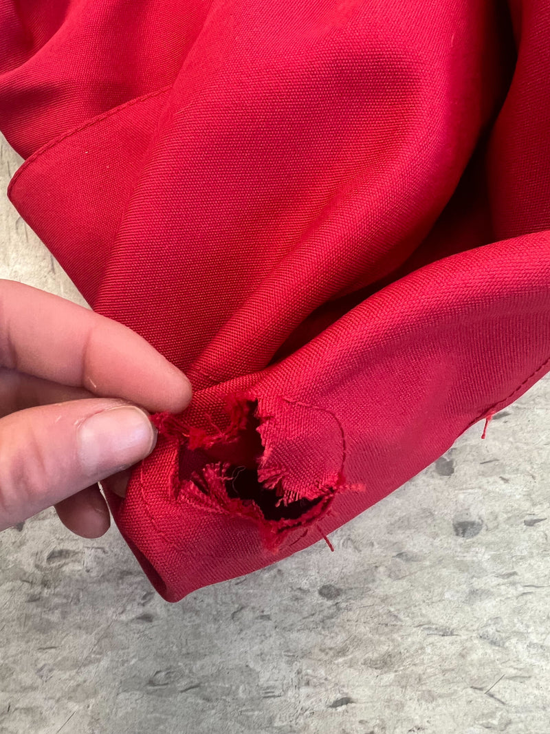 A person's hand is holding a red cloth with a significant tear. The cloth appears frayed and damaged, with threads visible around the hole. The background features a gray, speckled surface. This material could benefit from the Defective, Cut Finial Hole 9ft Market Patio Umbrella 6 Rib Replacement Canopy in Red by Formosa Covers to provide better coverage and protection.